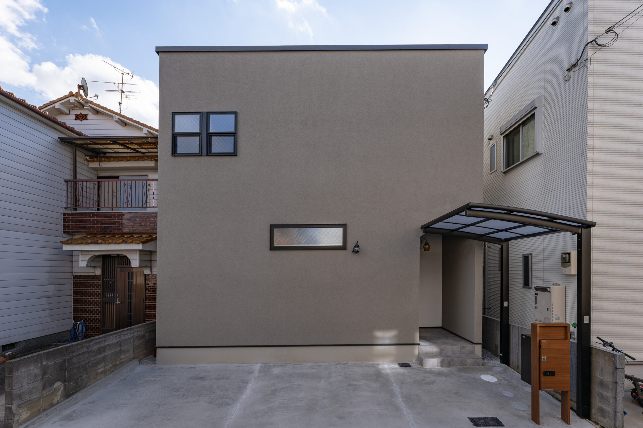 グレーのフラットな外壁に黒いサッシの小窓がアクセントになった、シンプルモダンなキューブ型住宅の正面外観。手前にはコンクリートの駐車スペースとカーポート、木目調の門柱を備えています。