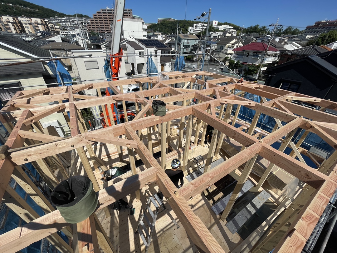 晴天の下で行われている木造住宅の上棟作業風景。クレーンを使用し、柱や梁、屋根を支える複雑な木構造がしっかりと組み上げられています。