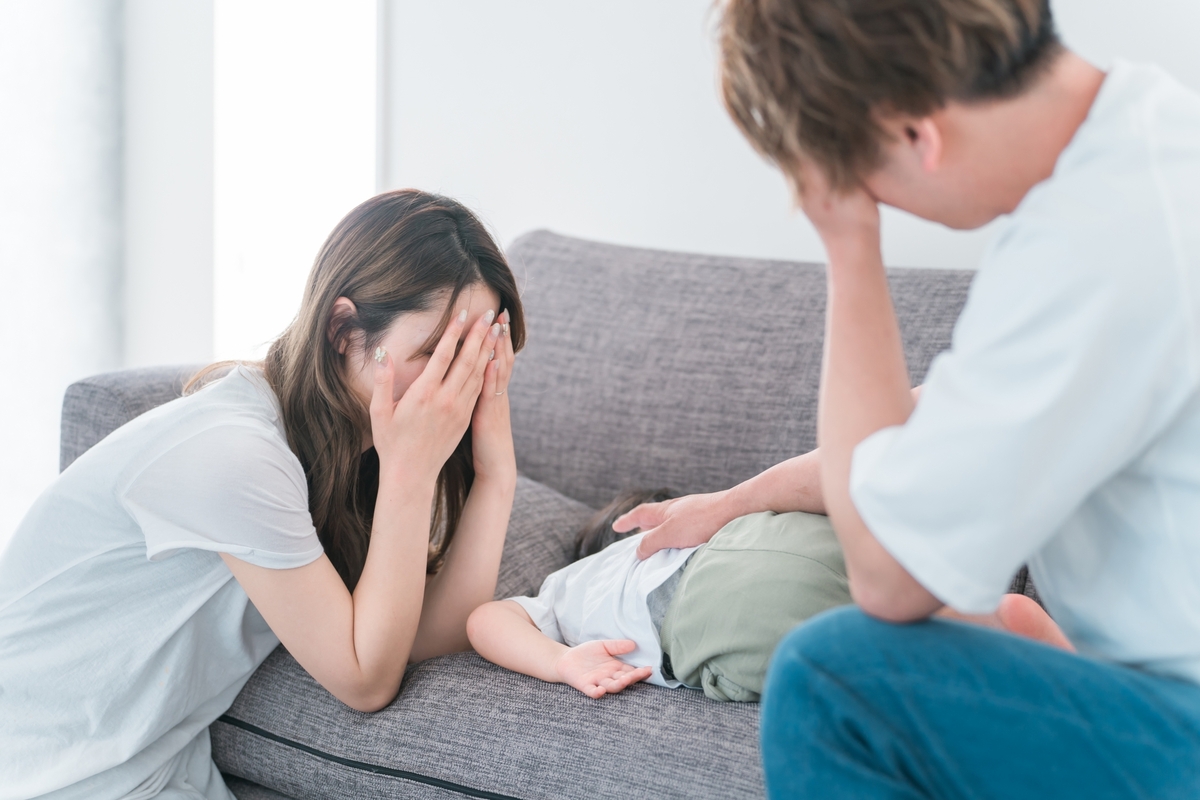 リビングのソファで頭を抱える男性と顔を覆う女性、横たわる子供。家づくりや住まいの悩みを感じさせます。