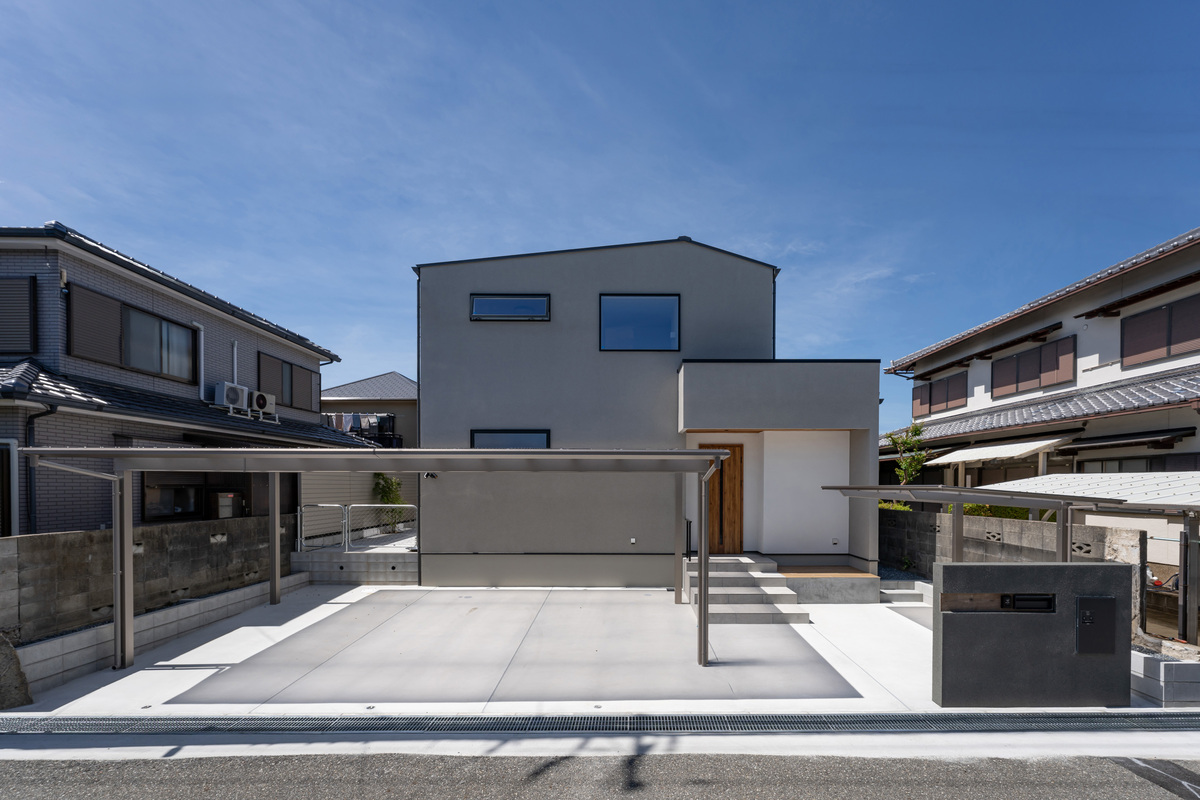 グレーの外壁と直線的なフォルムがスタイリッシュなモダンデザインの住宅外観。カーポート付きの広い駐車スペースを備えています。