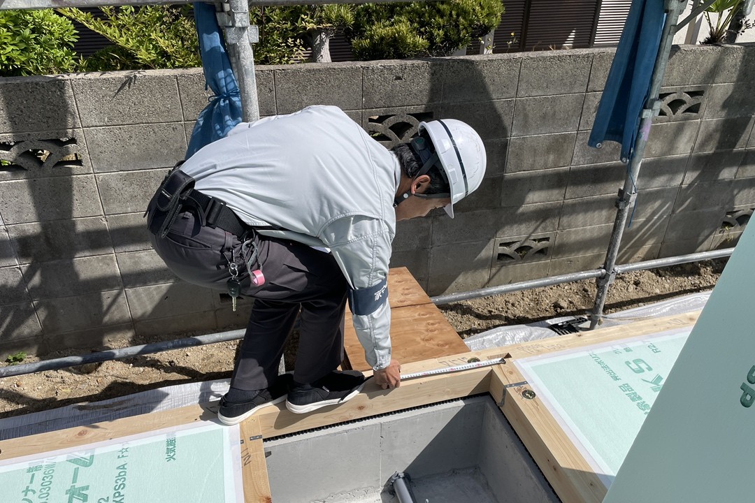 建築中の現場で、ヘルメットと作業着を着用した施工スタッフが、図面や基準を確認しながら基礎の施工精度を測定している様子。