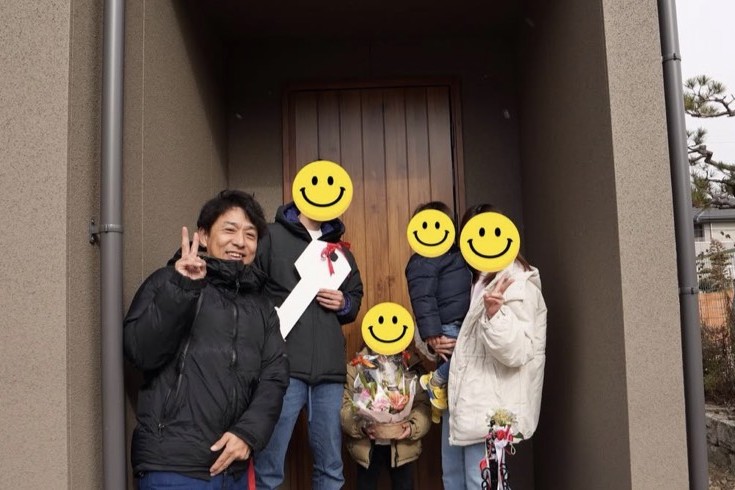 木目の玄関ドアとグレーの外壁が落ち着いた雰囲気の新居前で、大きな鍵のオブジェや花束を持ち、お引き渡しを祝うご家族と担当者。
