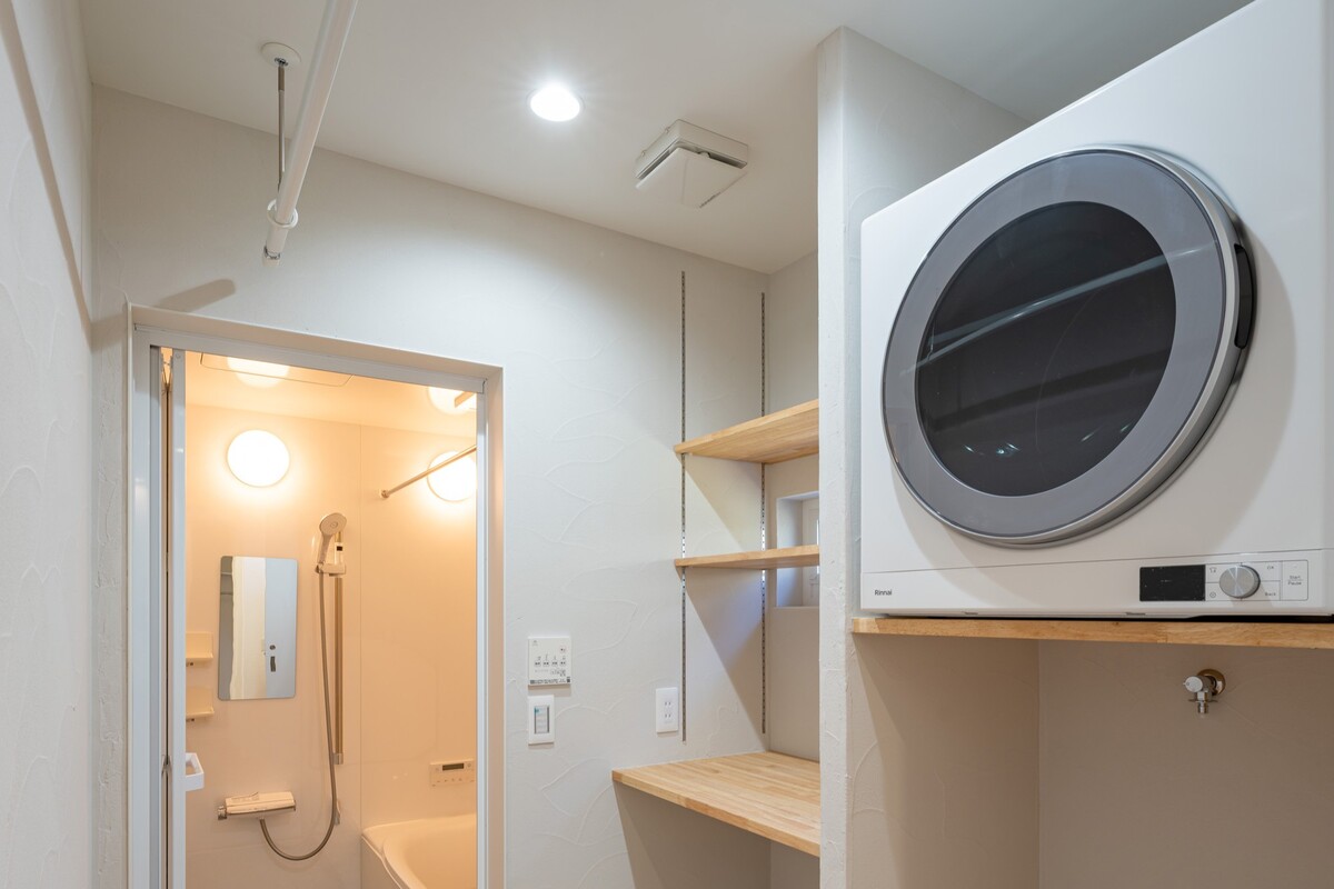 白を基調とした清潔感のある洗面脱衣室。造作の可動棚と木のカウンターの上に衣類乾燥機がすっきりと設置され、奥には明るいシステムバスが見えます。