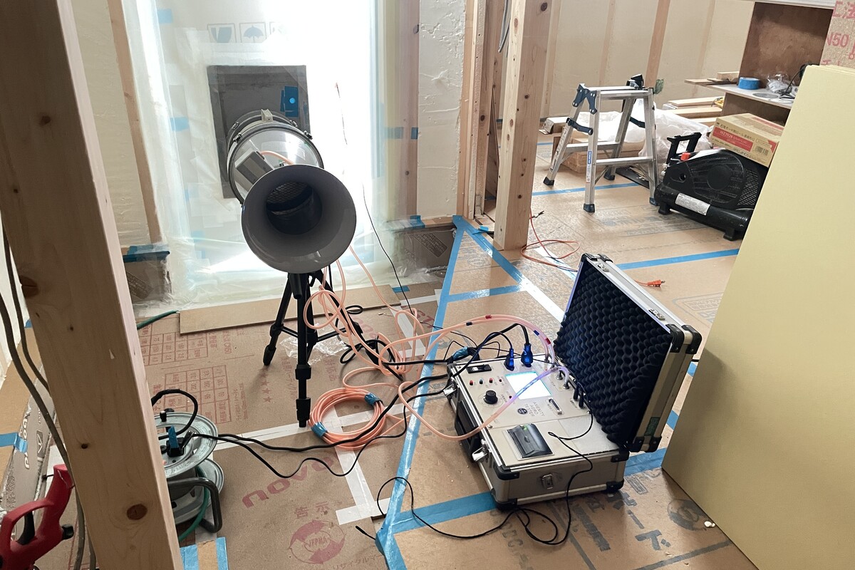 建築中の木造住宅の室内で、窓枠に専用の送風機を設置し、建物の隙間面積を計測する気密測定(C値測定)を行っている施工現場。