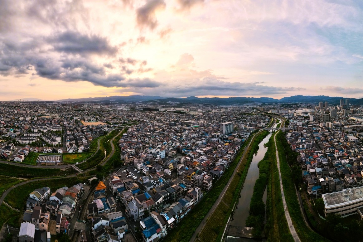 夕暮れの茜空が広がる街並みと、蛇行する川、低層の住宅が密集する高槻市周辺の広大な空撮風景。
