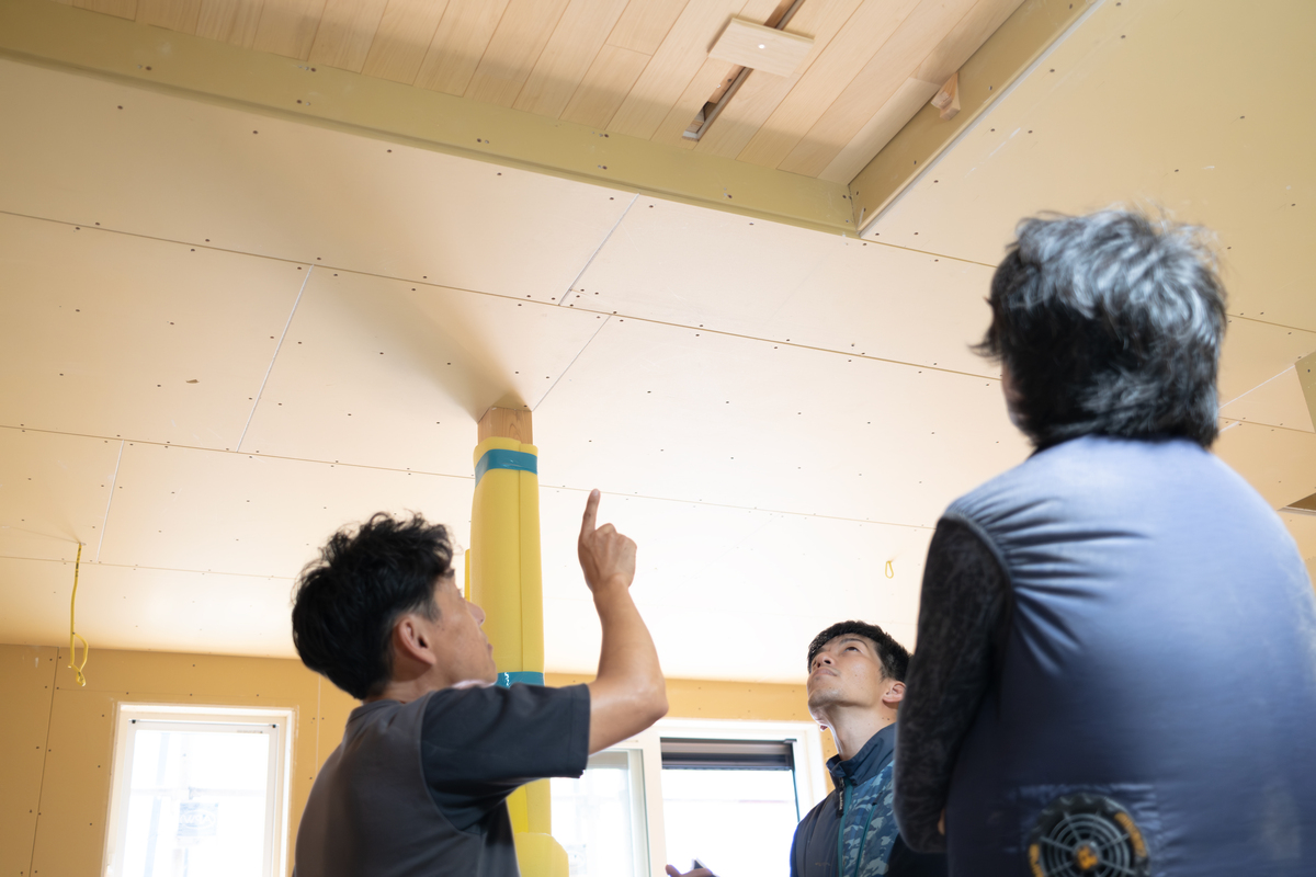 石膏ボードが張られた建築中の室内で、施工中の木製天井を指差しながら確認や打ち合わせを行う工務店スタッフと施主。