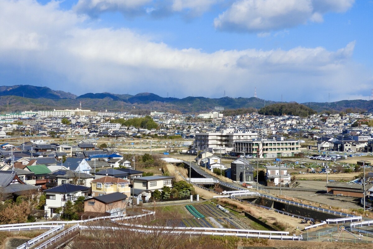 山並みを背景に、戸建て住宅や田畑が広がる穏やかで自然豊かな住宅街の風景。