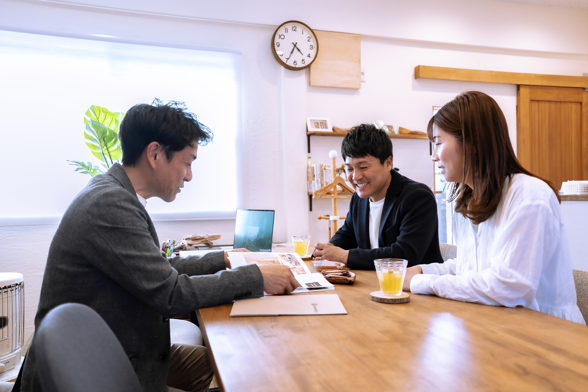 木の温もりを感じる無垢材のテーブルで、工務店スタッフとカタログを開きながら笑顔で家づくりの打ち合わせをしているご夫婦。