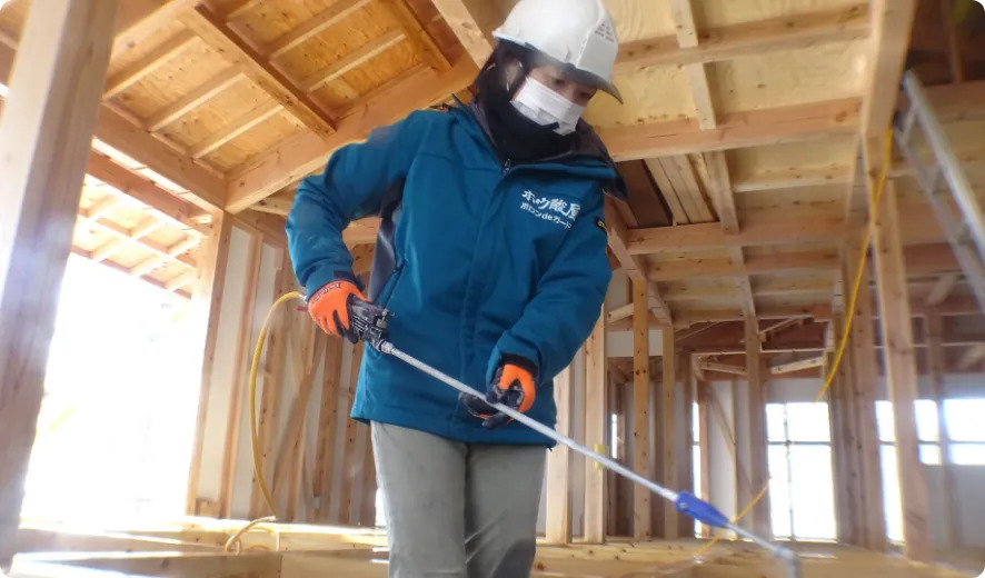 建築中の木造住宅で、ヘルメット姿の作業員が基礎や柱などの構造材にホウ酸による防蟻処理を行っている施工現場。