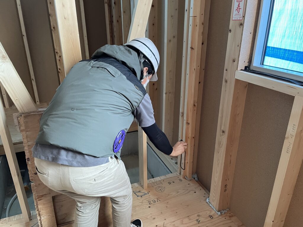 建築中の住宅内部で、ヘルメットと空調服を着用した現場監督が木造の柱や床下地枠の施工状態を細かく確認している様子。