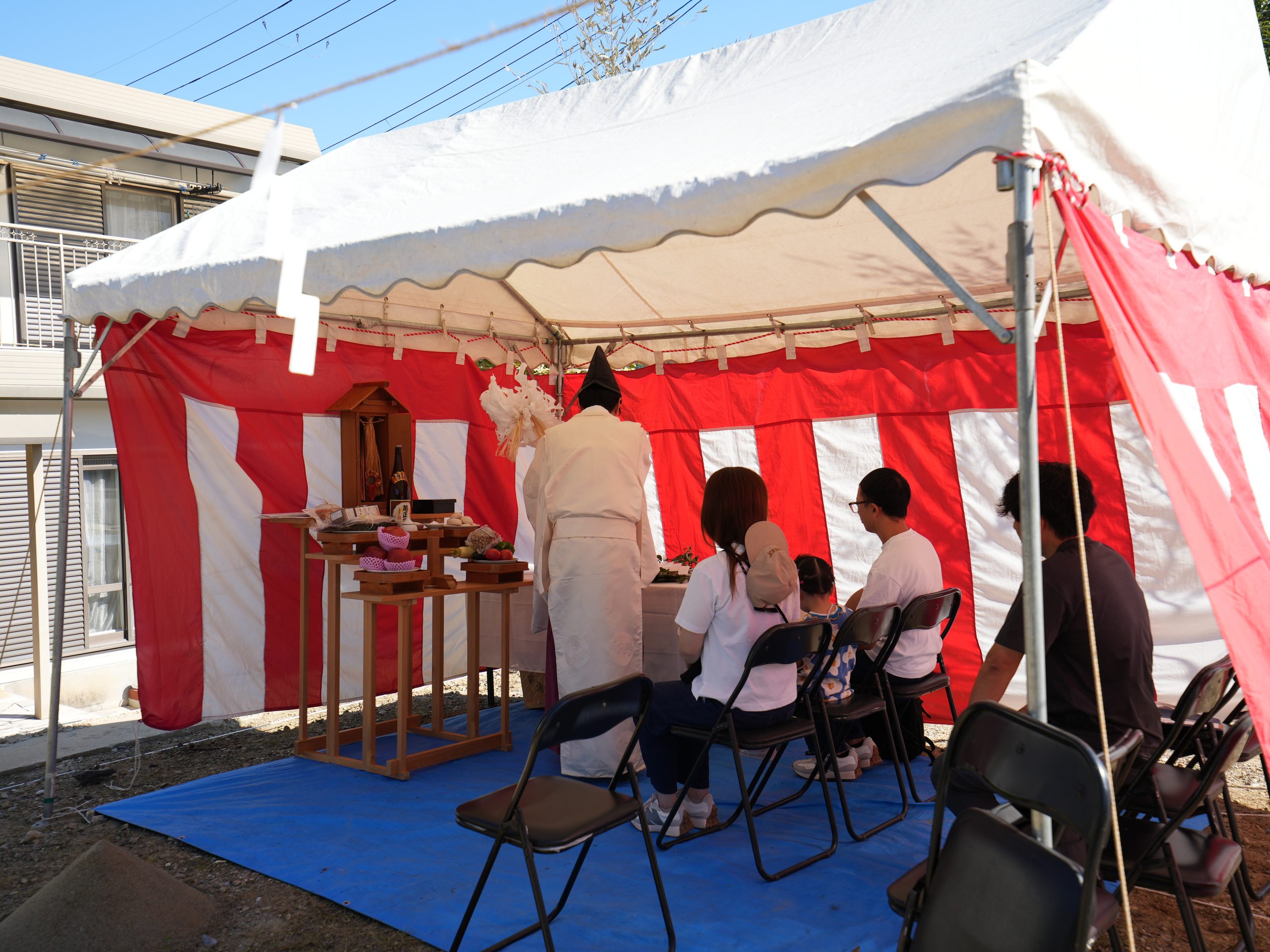 紅白幕を張ったテント内で執り行われている地鎮祭の様子。祭壇の前で神職が儀式を進め、施主様ご家族が着座して工事の安全を祈願しています。