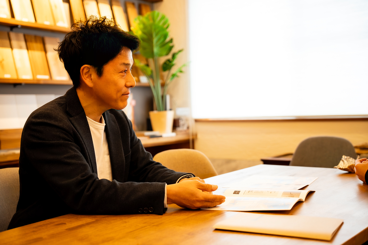 木材サンプルが並ぶ棚を背景に、無垢材のテーブルでカタログを開きながら笑顔で打ち合わせを行う工務店のスタッフ。