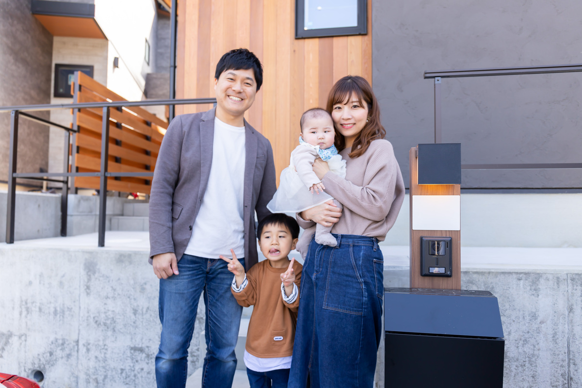 木の温もりを感じる縦張りの外壁とグレーの塗り壁が調和するモダンなマイホームの前に立ち、笑顔を見せる4人家族。