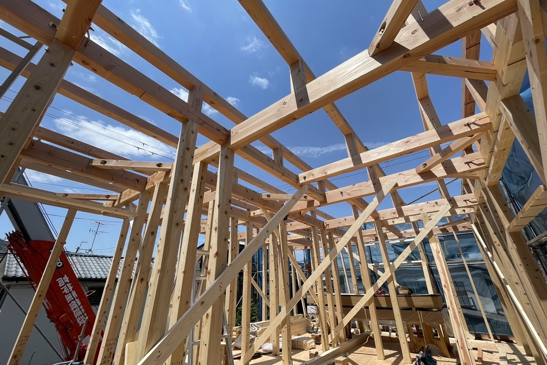 青空を背景に、新築住宅の木造軸組工法による頑丈な梁や柱が組まれた建て方工事中の躯体構造。