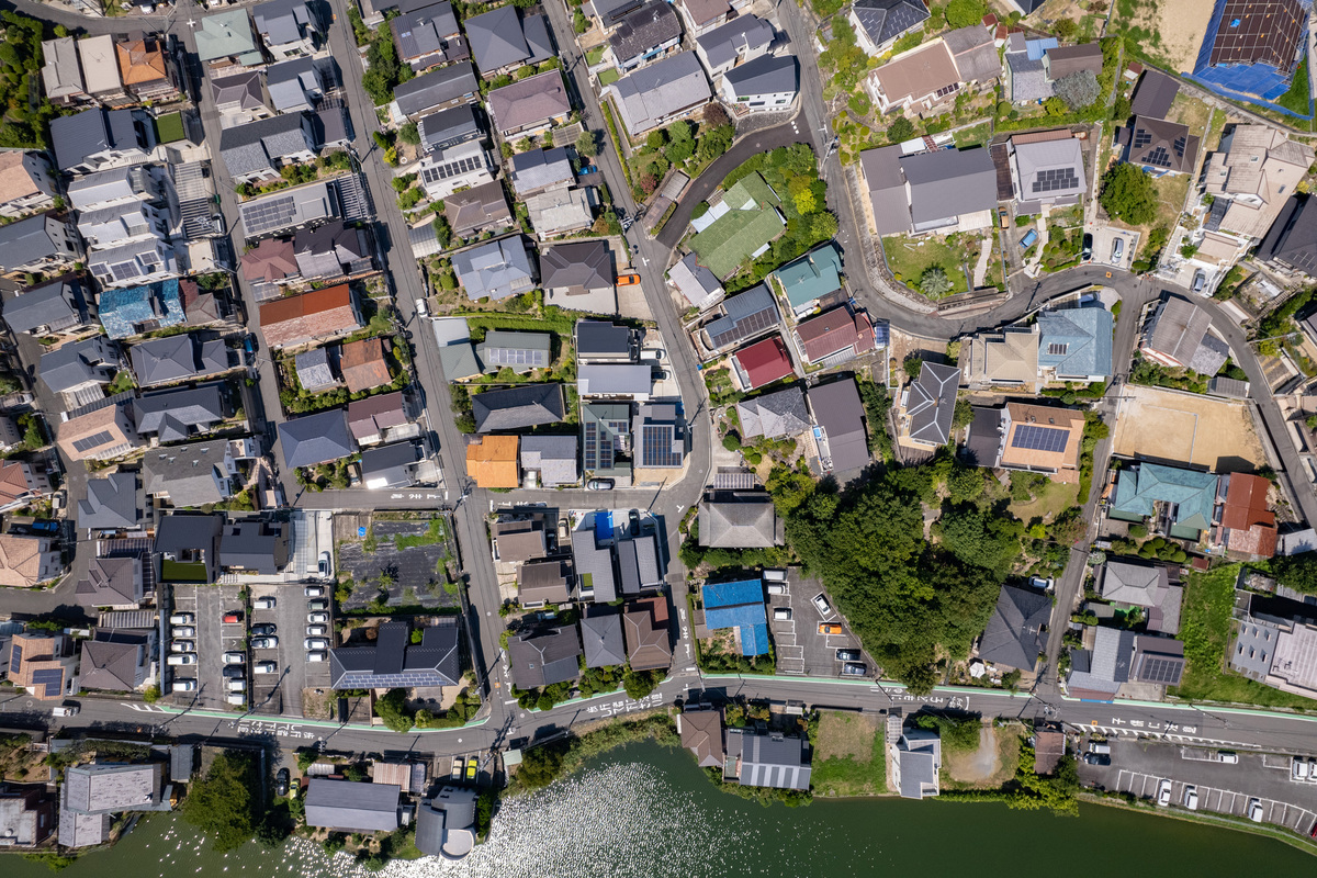 川沿いに区画整理された住宅街の俯瞰風景。様々な屋根形状の家屋や太陽光パネルが設置された住宅が密集して立ち並ぶ様子。