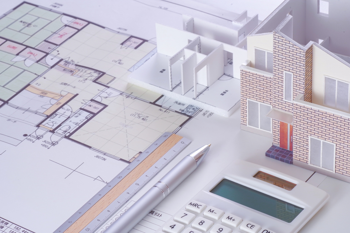 木造住宅の設計図面の上に配置された、外壁タイル調の2階建て住宅模型と室内間取り模型、三角スケール、電卓
