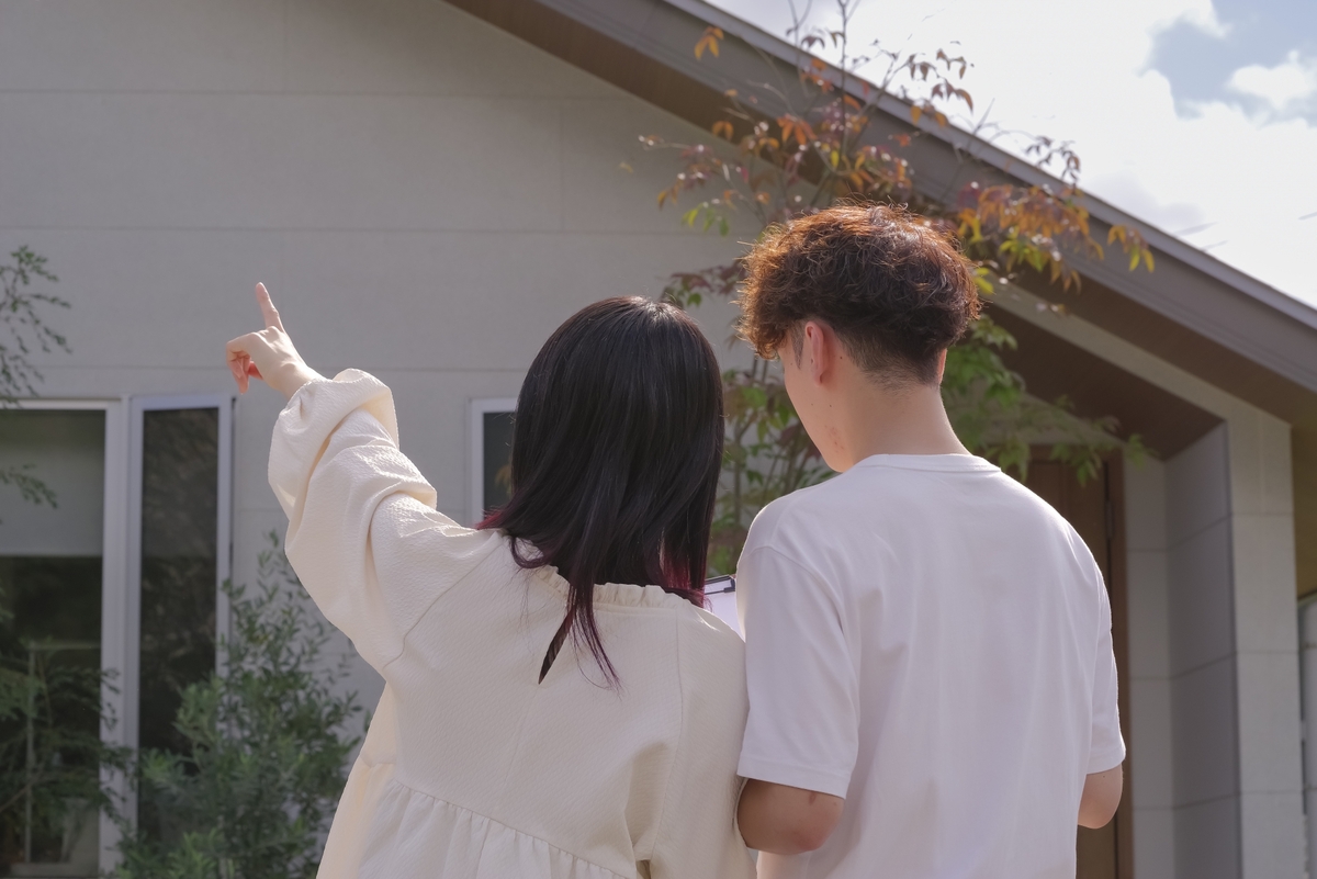 植栽が彩る明るい外壁の住宅の前で、バインダーを手に持ち、建物の外観や仕様を指差しながら確認している見学中のご夫婦の後ろ姿。