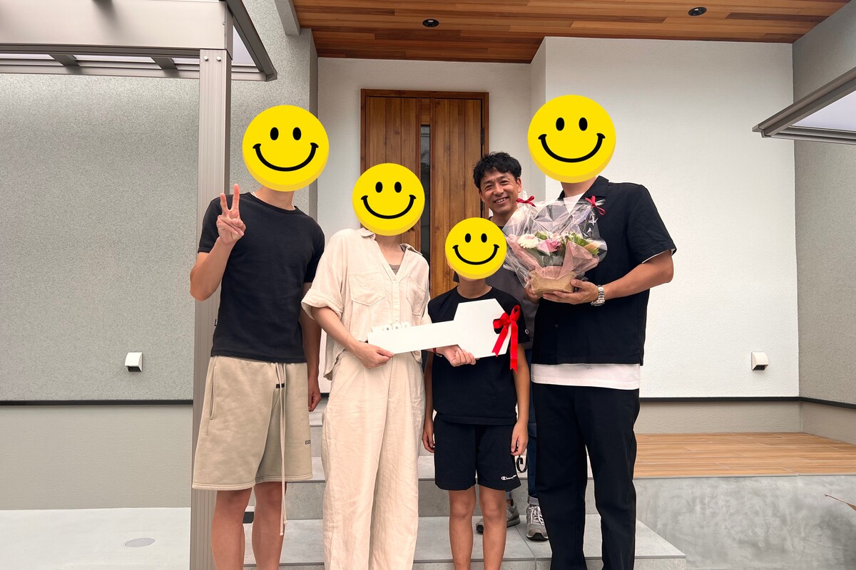 木目調の玄関ドアと軒天がアクセントになった新居の前で、大きな鍵の模型や花束を手に、引き渡しの記念撮影をするご家族と工務店スタッフ。