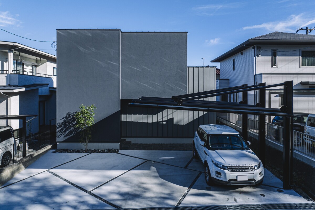 グレーの塗り壁と金属調の外壁を組み合わせたシンプルモダンな住宅外観。幾何学的な目地の土間コンクリートと黒いフレームのカーポートが設置された駐車スペース。