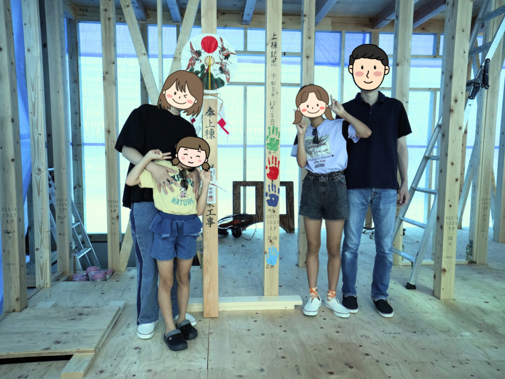 上棟と手形式を終えた建築現場で、お祝いの御幣やカラフルな家族の手形が記された構造柱を囲み、笑顔で記念撮影をする4人家族。木組みの梁や柱が露出した、家づくりの節目を感じる温かい光景