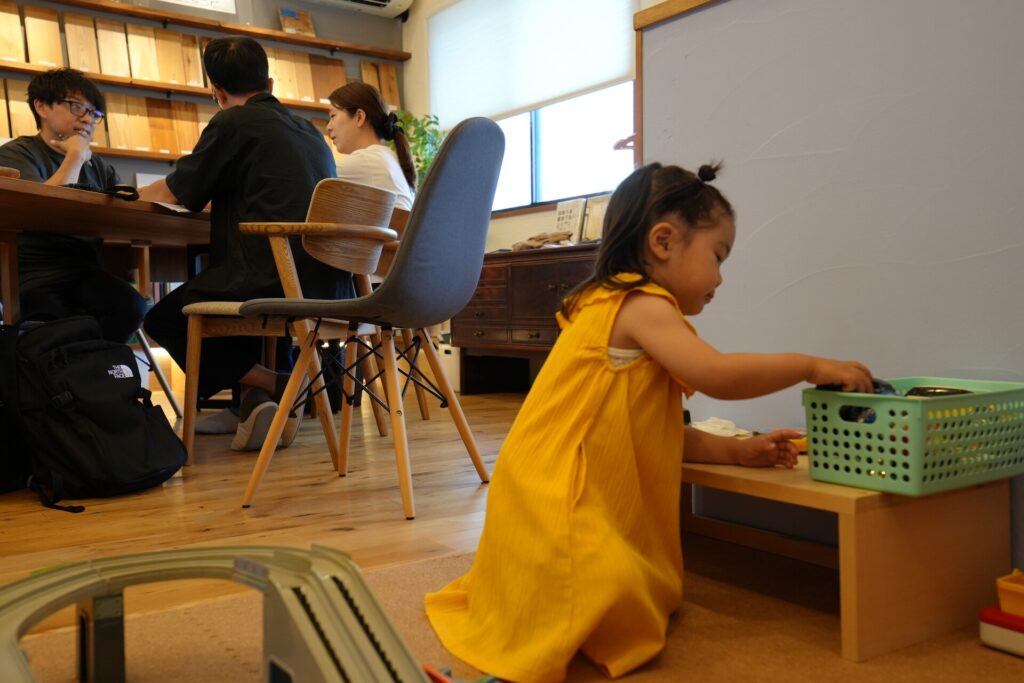 建材サンプルが並ぶ空間で大人たちが家づくりの打ち合わせをする傍ら、キッズスペースでおもちゃで遊ぶ子どもの様子。
