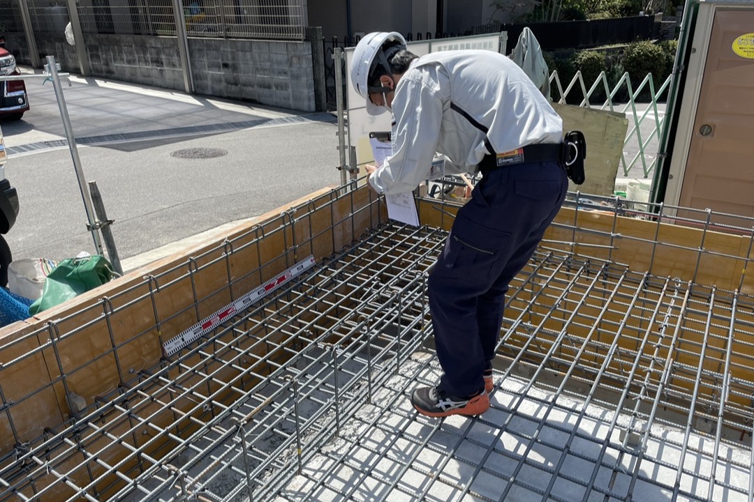 住宅の基礎工事現場で、組まれた鉄筋にスケールを当て、配筋の間隔や正確な施工状態を細かく確認・検査している作業員の様子。