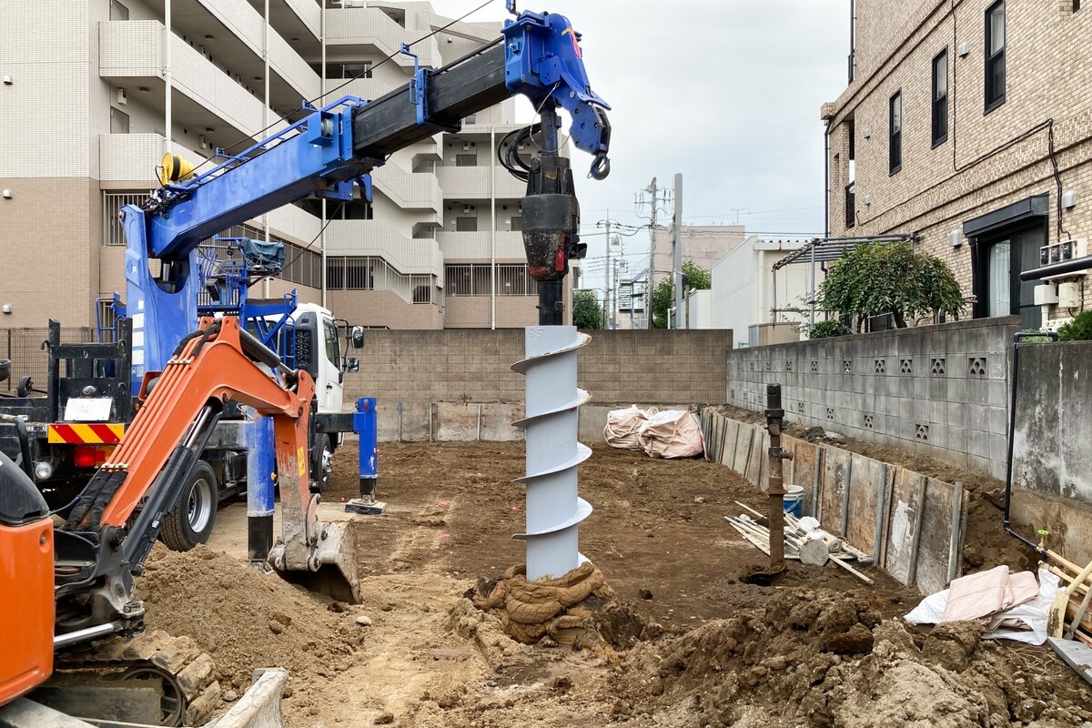 住宅建築現場において、青い重機のアースオーガーを用いて地面を掘削する地盤改良工事の様子