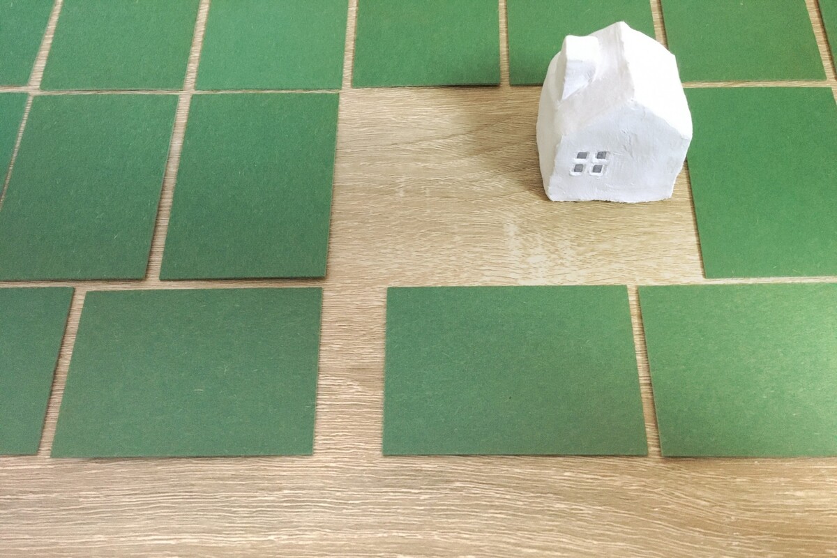 区画された緑のカードが並ぶ木目テーブルの上に置かれた、小さな白い家型オブジェ