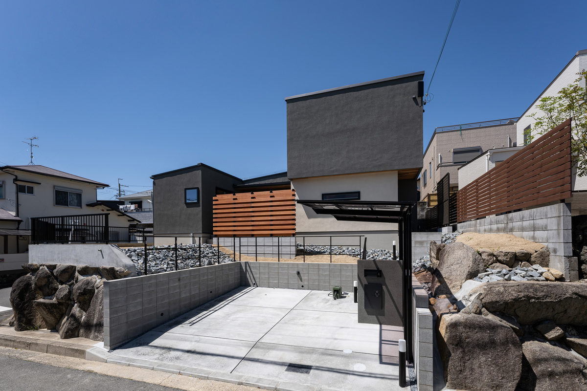 グレーと白のツートンカラーの外壁に木目調のルーバーがアクセントになったモダンな住宅の外観。高低差のある敷地を活かした石積みと駐車スペースが特徴です。