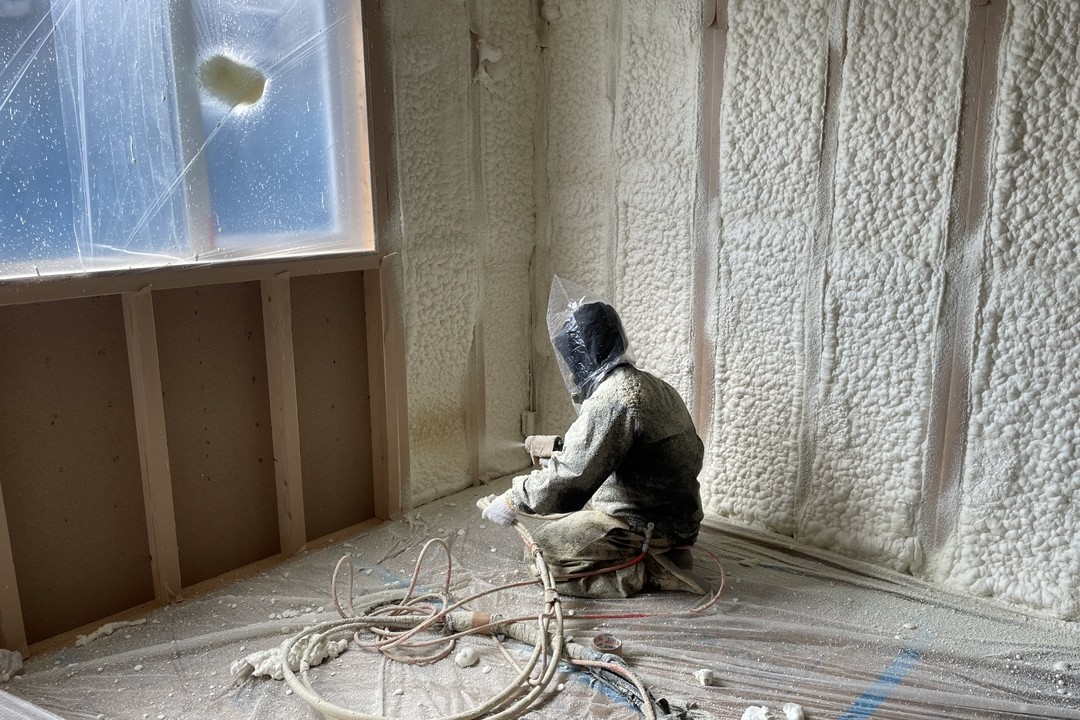 建築中の住宅内部で、防護服を着た作業員が壁一面に発泡ウレタン断熱材を吹き付け施工している様子。