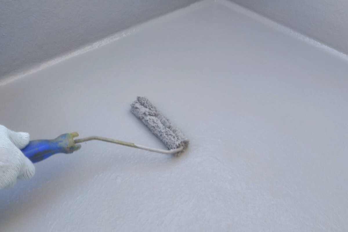 バルコニーの床面に、作業員がローラーを使ってグレーの防水塗料を丁寧にムラなく塗布している防水工事の施工風景。