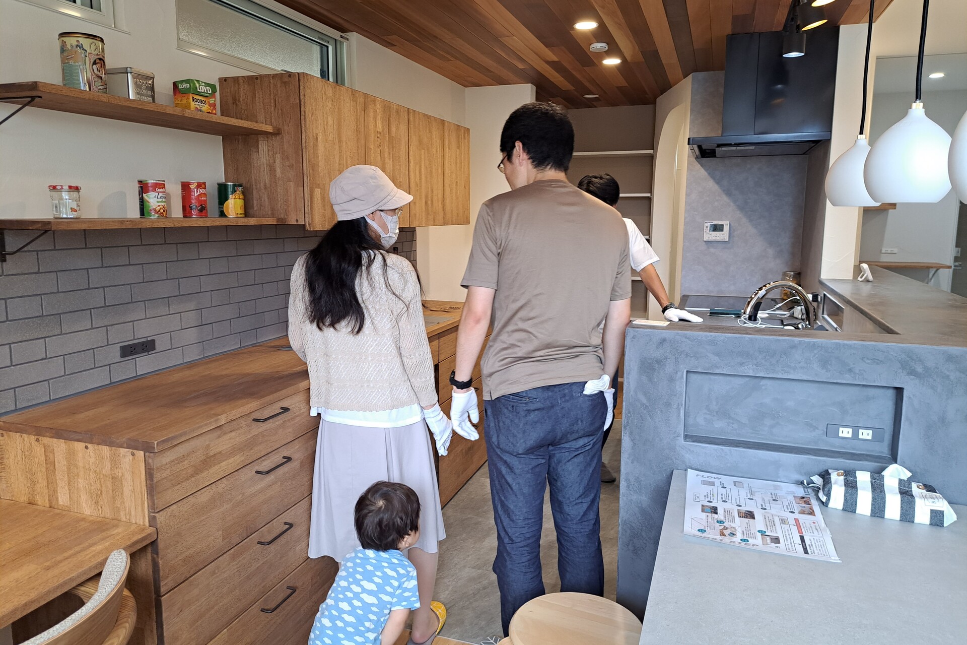 板張りの天井やモルタル調のカウンターなど、素材感にこだわったキッチンスペースを見学する家族。